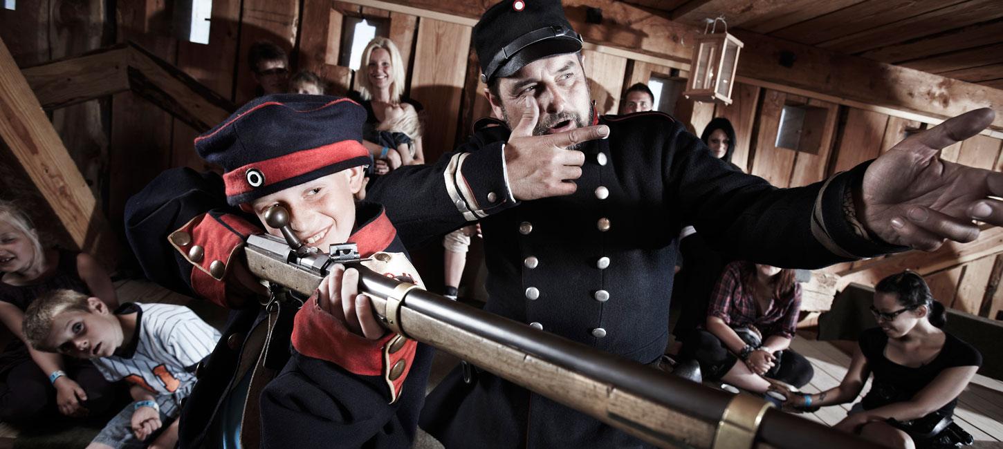 Medarbejder (formidler) og barn i uniform skyder med trægevær på Historiecenter Dybbøl Banke